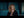 A blonde woman in a black top looks directly at the camera against a dark blue-toned blurred background. White text in the bottom right reads 'MG RED Log3G10 V2 Warm'.