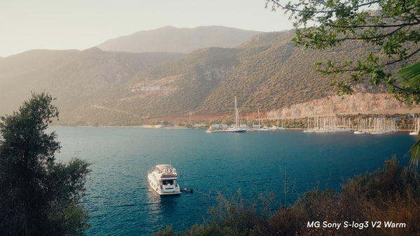 A scenic coastal landscape with deep blue water, a white motorboat in the foreground, sailboats at a harbor, and mountains covered in green vegetation under warm golden-hour light. Tree branches frame the top right corner, and a text watermark in the bottom right reads 'MG Sony S-log3 V2 Warm'.