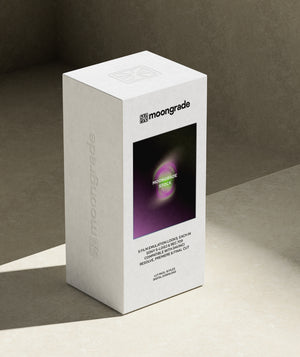 A white Moongrade cosmetic box standing upright on a beige surface against a textured gray background. The front of the box features a black square window showing a purple and green circular graphic with the text 'MOONGRADE STOCK